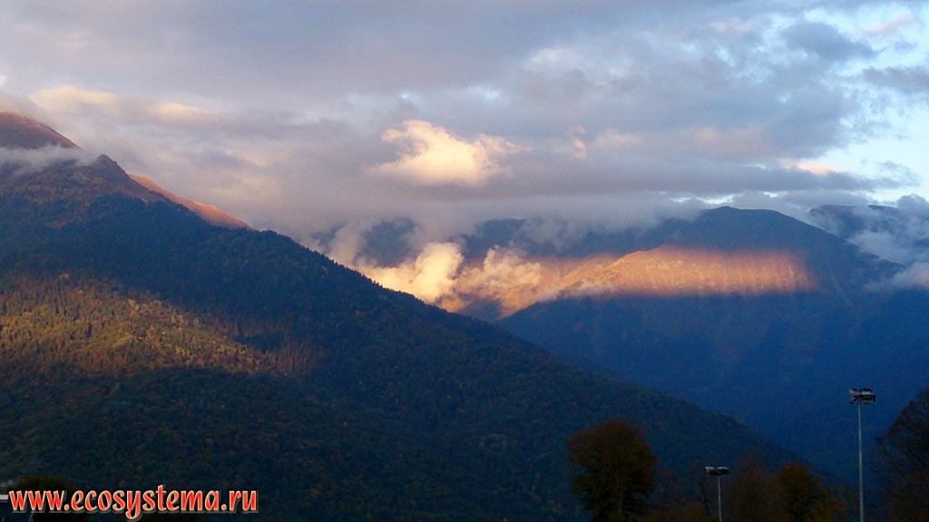 Лучи солнца, пробивающиеся сквозь дождевые облака и освещающие горные хребты на территории Кавказского государственного биосферного заповедника, покрытые широколиственными лесами на закате после дождя