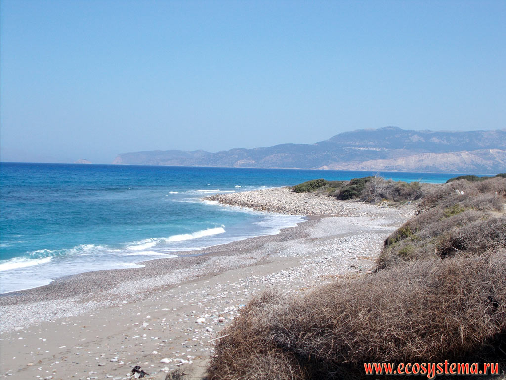 Песчано-галечный пляж и голубые воды Эгейского моря на северо-западном побережье острова Родос со средневысотными горами, сложенными известняками