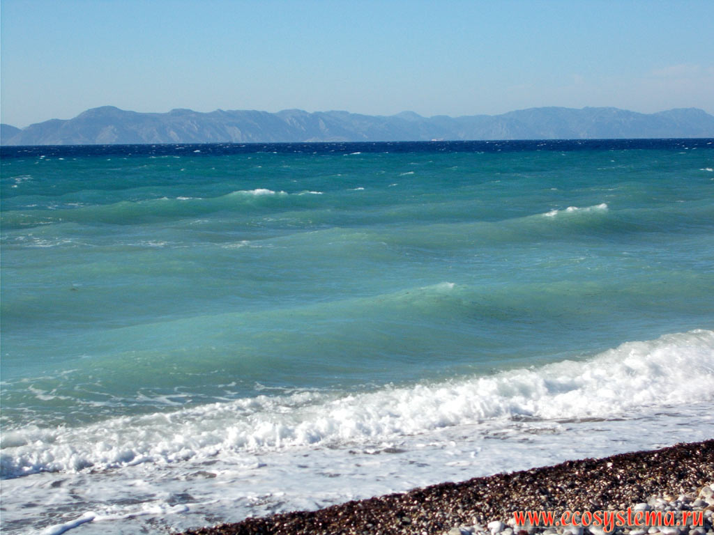Прибой на галечном пляже, голубые воды Эгейского моря и вид на горы Тавр и полуостров Мармарис в Турции на северо-западном побережье острова Родос